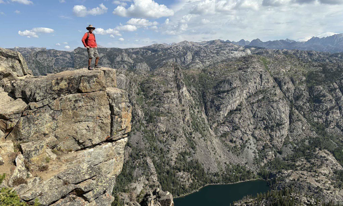 A man in jeans, red shirt, and a hiking hat stands on a rocky point.